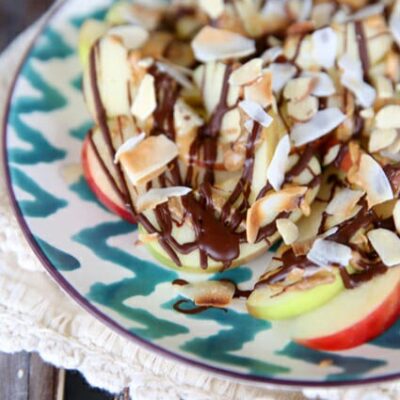 A teal and white plate of apple slices, covered in a drizzle of peanut butter and chocolate, topped with coconut and almonds.