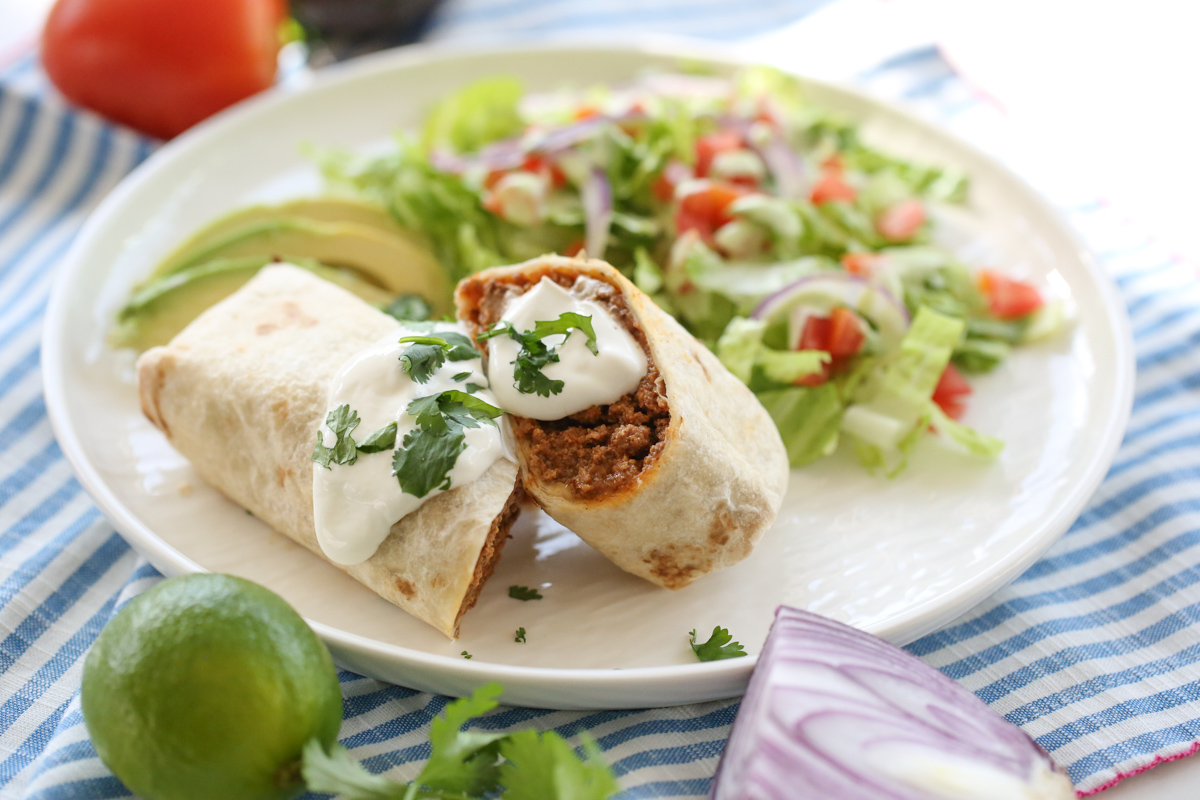 burrito on a white plate cut in half stuffed with bean, beef, and cheese. 